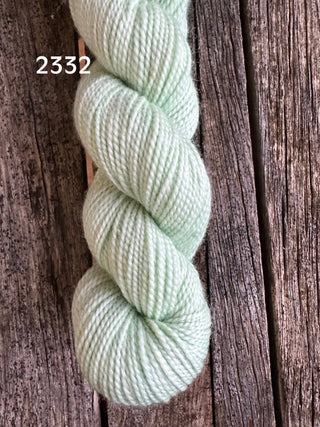 A skein of Koigu KPM & KPPPM Fingering Weight Yarn in pale mint green, labeled 2332, sits on a weathered wooden surface, showcasing the rustic cracks and texture beneath.