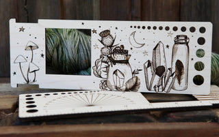 The Apothecary "Almost Everything" Ruler is a wooden yarn measuring tool engraved with mushrooms, crystals, a jar, stars, and a crescent moon. Yarn shows through its large window. The ruler rests on a rustic wood surface.