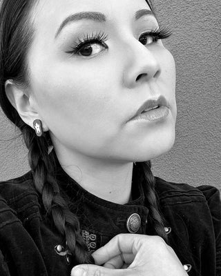 Black and white close-up of a person with braided hair, makeup, and handmade Wednesday Stud Earrings, hypoallergenic and cat-shaped from San Francisco, looking confidently at the camera against a textured background. Their hand rests near their collar.
