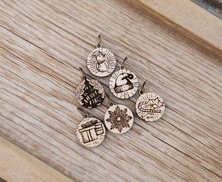 The Maple Wood Yuletide Knitting Stitch Markers Set of 6 features festive designs—snowman, polar bear, Merry Xmas tree, cat in a box, snowflake, and wrapped gift—displayed on a rustic wooden surface.