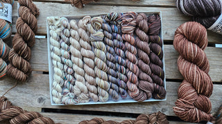A Koigu Pencil Box in Mocha Mousse, filled with soft Merino wool fingering weight yarn in cream to dark brown gradients, sits on a wooden surface with additional brown-toned skeins nearby.