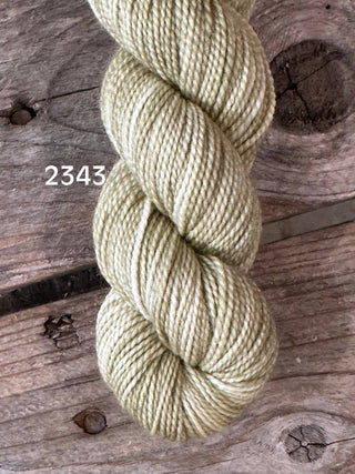 A skein of Koigu KPM & KPPPM Fingering Weight Yarn in light olive green, color 2343, sits on a rustic weathered wooden surface.