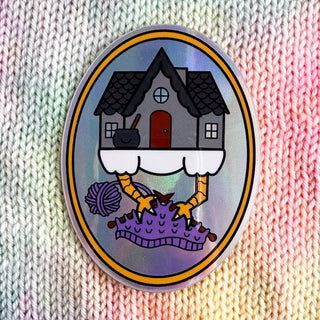The Baba Yaga Knitting Holographic Sticker features a house with chicken legs on clouds, knitting a purple project against a rainbow background—adding witchy craft charm to any collection.
