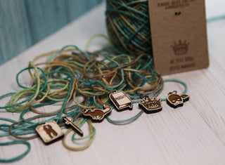A ball of multicolored yarn rests on a light surface. In front, six Princess Bride Knitting Stitch Markers—maple wood accessories shaped like a cat, sword, dinosaur, book, campfire, and mask—are neatly lined up for display. A brown paper tag is partially visible in the background.