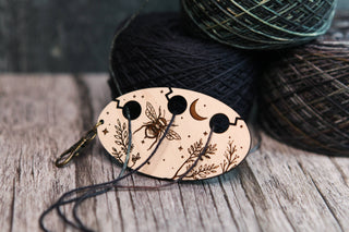 The Yarn Management Tool for Knitters features engraved designs of a bee, leaves, moon, and stars, and holds three strands of dark thread. Ideal as a knitting tool or crochet accessory, it is pictured beside two balls of dark yarn on a rustic wooden surface.