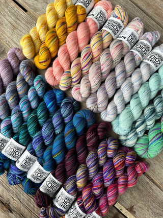 Skeins of Bridgerton Collection KPPPM yarn are arranged in rows on a wooden surface, showcasing a beautiful gradient of colors including yellows, pinks, neutrals, blues, greens, purples, and reds.