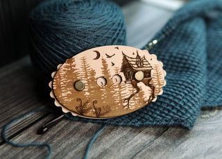 The Baba Yaga Row Counter, featuring a wooden pin with a forest and house design, rests on blue knitted fabric beside a ball of yarn and a crochet hook on a wooden surface.