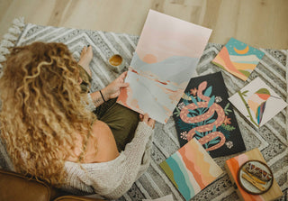 A person with curly hair sits on a patterned rug, holding a pastel landscape from the Walk in Nature Meditative Art Paint by Number Kit, surrounded by colorful abstract paintings—a soothing creative ritual with coffee and art supplies nearby.