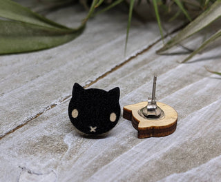 🐈‍⬛ Black Cat Earrings—small black cat shapes with white eyes and mouth—are displayed with their wooden and metal backs on a light surface and green leaves, making cute hypoallergenic earrings perfect for Halloween.