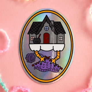 The Baba Yaga Knitting Holographic Sticker shows her house with chicken legs holding knitting needles and yarn balls on a pink, fuzzy background for cozy witchy charm.