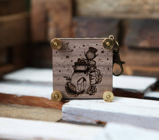 The Miniature Flower Press is a small wooden keychain engraved with a jar of thistle flowers, stars, and a crescent moon, secured with brass fasteners and displayed on stacked wooden planks—a pocket-sized floral keepsake.