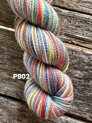 A skein from the KPM and KPPPM Variegated Color Collection features red, blue, yellow, green, and gray hues twisted together on rustic wood. P802 appears on the left—perfect for hand knitting projects.