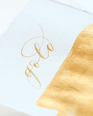 A white paper showcases gold written in elegant gold calligraphy using Premium Calligraphy Ink, beside a brushstroke of shimmering gold paint.