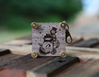 The Miniature Flower Press is a small wooden keychain with a metal clasp, engraved with a jar, flowers, leaves, stars, and a crescent moon—a pocket-sized charm that stands upright on outdoor surfaces.