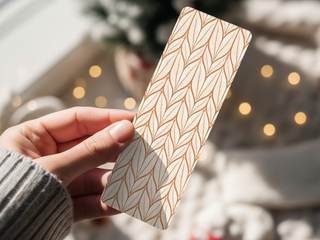 A hand holds a rectangular Holiday Bookmarks for Knitters printable bookmark featuring a gold leaf pattern, set against a softly blurred background with warm lights and touches of greenery.