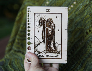 A hand holds ✨ The Hermit ✨ Tarot Knitting Needle Gauge & Ruler, crafted from maple wood, featuring measurement markings and gauge holes along its edges. It rests on green knitted fabric.