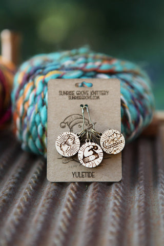 A set of six maple wood Yuletide knitting stitch markers with festive designs is displayed on a labeled card, seen in front of colorful bulky yarn skeins—perfect for celebrating the Christmas holiday season.