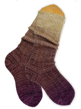 A pair of gradient socks, made from Solemate Sock Yarn, is displayed on a wooden sock blocker, featuring a smooth fade from beige at the cuff to deep purple at the toes.