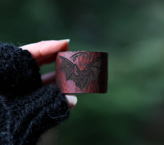 A hand in a black fingerless glove holds the 🦇 Vampire Bat Under the Moon Shawl Cuff 🦇, a handcrafted leather accessory with an engraved bat design—ideal as a unique shawl pin or for knitting and crochet, set against a blurred green background.