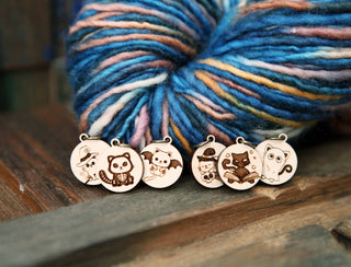 The 🐾 Scaredy Cats Stitch Marker Set 🐾 features five maple wood stitch markers with cute Halloween cat engravings, displayed in front of a colorful skein of yarn—perfect festive accessories for your knitting projects.
