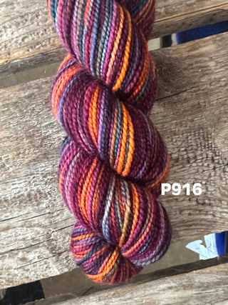 A skein from the KPM and KPPPM Variegated Color Collection in color P916, featuring burgundy, orange, purple, and gray hues, rests on rustic wood. Ideal for hand knitting projects. The P916 label is displayed beside the soft merino yarn.