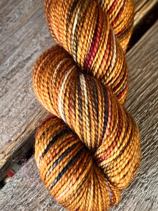 A close-up of a KPM and KPPPM Variegated Color Collection Skein in gold, brown, black, cream, and red rests on rustic wood—ideal for hand knitting projects.