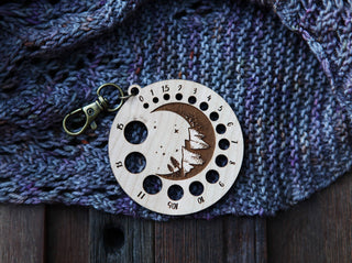 The Crescent Moon Knitting Needle Gauge, a round cherry wood keychain engraved with mountains, stars, and a crescent moon, shows numbered hole sizes as it rests on purple knitted fabric.