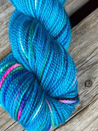 A close-up of a skein from the KPM and KPPPM Variegated Color Collection shows bright blue yarn with pink, green, and purple flecks on weathered wood—ideal for hand knitting projects.