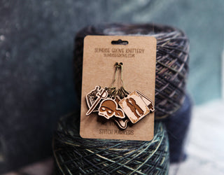 A set of Princess Bride Knitting Stitch Markers, crafted from metal and maple wood, displayed on a branded card atop two skeins of multicolored yarn with a blurred background—a charming addition to your knitting accessories collection.
