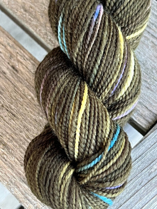 A close-up of a KPM and KPPPM Variegated Color Collection Skein in tightly twisted olive green with pastel streaks of yellow, blue, and purple, resting on weathered wood—ideal for hand knitting projects.