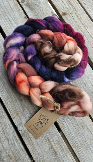A Koigu Merino Roving Braid in purple, red, pink, orange, and brown rests on wood. A kraft tag with a tiger illustration is attached with twine—ideal for spinning fiber projects.