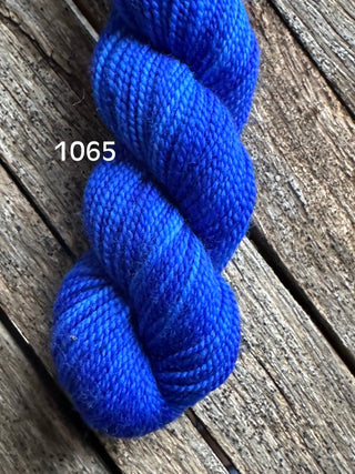 A skein of Koigu KPM & KPPPM Fingering Weight Yarn in bright blue, labeled 1065, sits on a rustic wooden surface with visible grain—ideal for knitting or crochet projects.