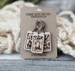Set of three wooden Tarot Card Stitch Markers—The Tower, The Devil & Judgement—feature intricate designs and come attached to a Sunrise Grove Knittery card, displayed against softly blurred cream-colored yarn.