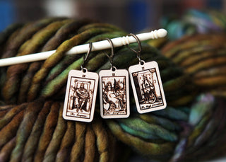 The Tarot Card Stitch Markers set features The High Priestess, The Empress, and The Emperor in maple wood—perfect progress keepers for your knitting or crochet projects. Shown on a white needle with vibrant yarn in the background.