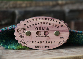 A wooden Ouija Board Row Counter with engraved letters, numbers, YES, NO, and GOOD BYE rests on a colorful knitted surface.