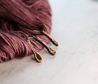 Close-up of 🧹 Witch’s Broom Stitch Markers 🧹 in burgundy yarn, ideal for knitting or crochet projects, displayed on a light, textured surface.