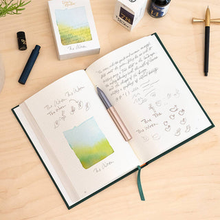 A journal open to a page of notes, doodles, and watercolor art is surrounded by The Wren - Refillable Writing Pen with a Japanese-made tip, an ink bottle, pen case, and card box on a light wooden desk.