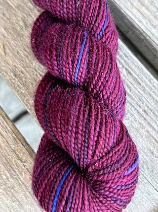 A skein from the KPM and KPPPM Variegated Color Collection glows in deep magenta with blue and purple accents, coiled on rustic wood—ideal for hand knitting projects.