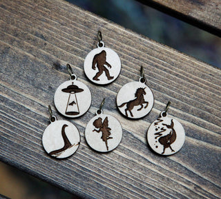The 👽 I Believe Stitch Marker Set 👽 includes six round maple wood markers engraved with silhouettes of knitting accessories and mythical creatures: UFO, Bigfoot, unicorn, Loch Ness Monster, fairy, and jackalope.