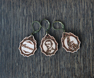 Three Nevermore Knitting & Crochet Stitch Markers, featuring engraved designs—Nocturne, a portrait, and sheet music—on wooden earrings with vintage-style metal hooks, are displayed on a dark wooden surface.