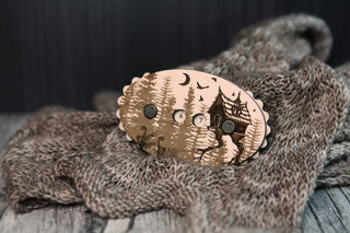 The Baba Yaga Row Counter, a maple wood brooch adorned with a whimsical forest cabin design and numbered buttons, rests on gray knitted fabric, set against a rustic wooden background.