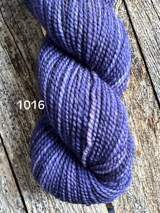 A skein of Koigu KPM & KPPPM Fingering Weight Yarn in deep purple, showing subtle shade variations, rests on weathered wood with the number 1016 displayed on the left side.