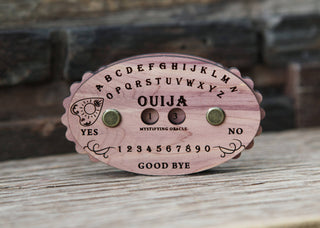 The Ouija Board Row Counter is a small wooden device featuring letters, numbers, “YES,” “NO,” and “GOOD BYE” inscribed, with two metal pegs and a circular window on the front, all set against a rustic wooden background.