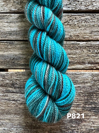 A skein from the KPM and KPPPM Variegated Color Collection, in turquoise, brown, white, and gray Koigu Premium Merino yarn, rests on a rustic wooden surface. Code P821 appears in white text at the bottom right corner.