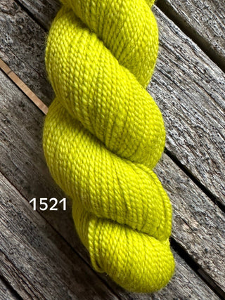 A skein of Koigu KPM & KPPPM Fingering Weight Yarn in bright yellow (label 1521) rests on a weathered wooden surface, its vibrant color standing out against the grain and gaps between the planks.