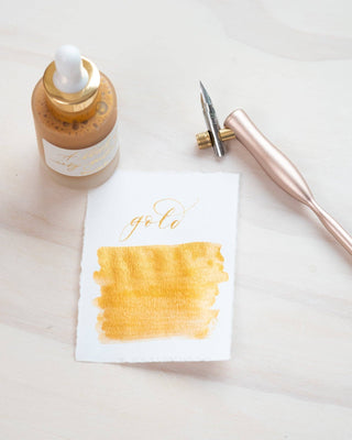 A bottle of Premium Calligraphy Ink, a metallic-handled dip pen, and a card with “gold” calligraphy above an ink swatch are displayed on a light wooden surface.