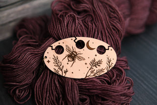 The Yarn Management Tool for Knitters is an oval wooden accessory, engraved with a bee, moon, stars, and leaves. Designed with three holes and intricate decorative patterns, this charming tool rests on dark maroon yarn—making it a delightful addition to any crochet or knitting project.