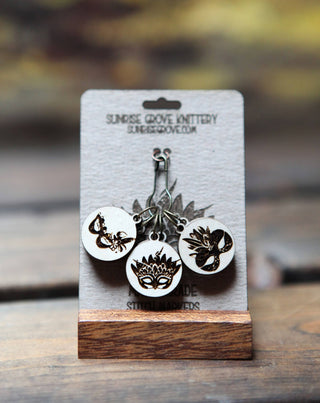 Six round maple wood stitch markers with laser-etched masquerade mask designs are displayed on a card labeled "Sunrise Grove Jottery," set on a wooden stand with a blurred Mardi Gras background. Product: Maple Wood Masquerade Mask Stitch Markers Set of 6.