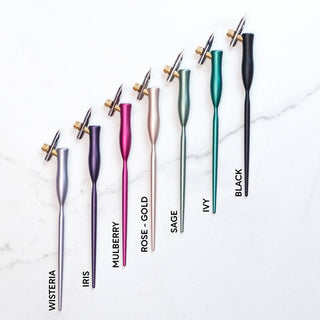 Seven Flourish Calligraphy Pens in wisteria, iris, mulberry, rose-gold, sage, ivy, and black with gold flanges are vertically lined up on a white surface—ideal for beginners.