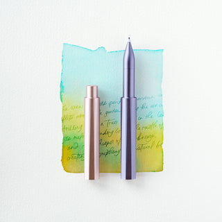 The Wren - Refillable Writing Pen and its cap are shown on a watercolor background with handwritten green ink, blending blue, turquoise, yellow, and green shades.